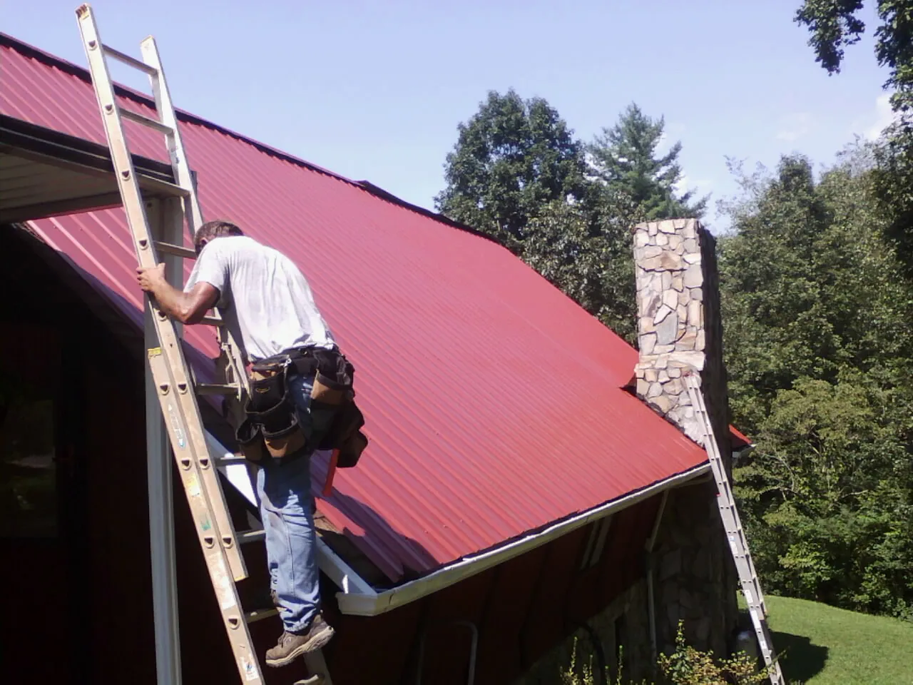 Roofer on a ladder beside a red metal roof and stone chimney — Soffit & Fascia Repair by Roofing Brothers in Willow Springs